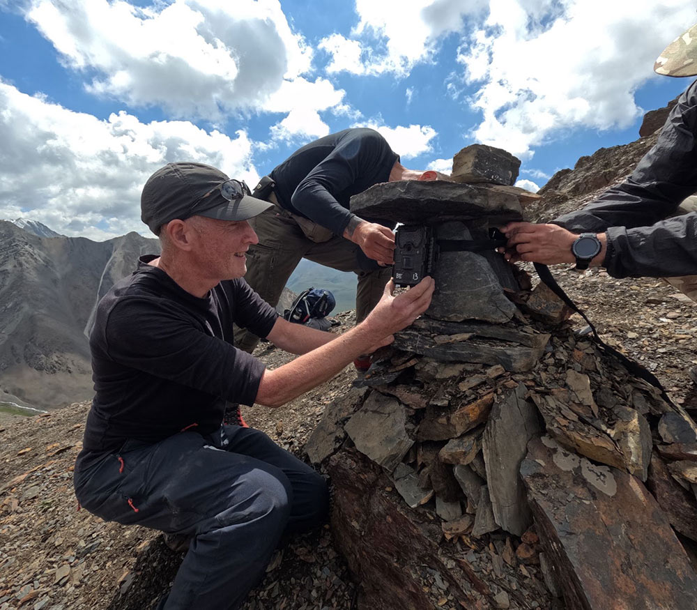 3 people around a pile of slate and rocks, setting up a camera trap, with a dry, mountainous landscape in the background